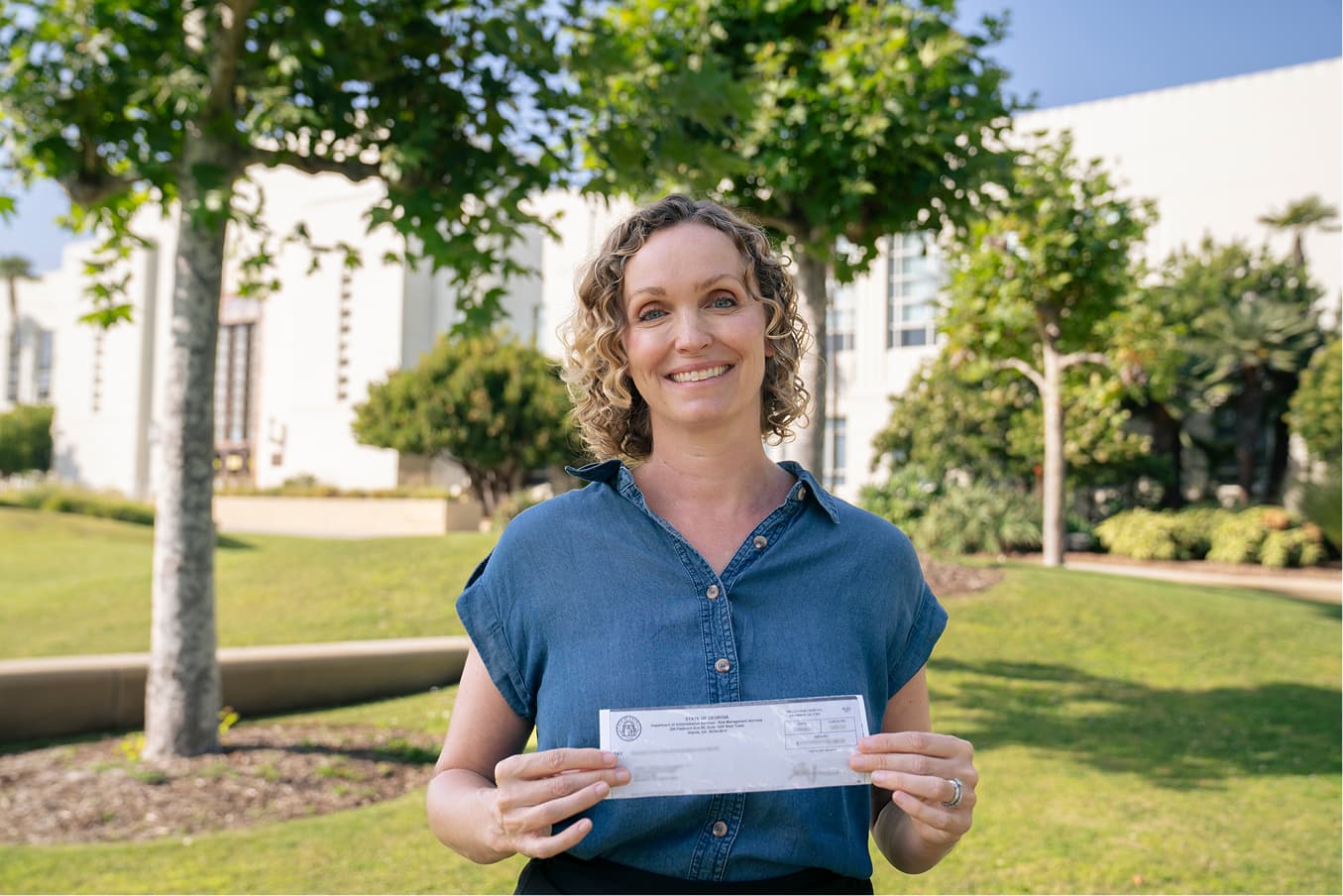 Smiling woman standing on a lawn holding up a state-issued check